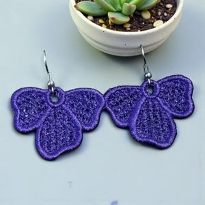 Op de afbeelding: Een paar paarse geborduurde oorbellen in een bloemmotief. De oorbellen zijn gemaakt van een textiel met textuur en hebben een donkerpaarse omtrek. De oorbellen hangen aan zilverkleurige oorhaken. Een kleine witte pot met een vetplant staat op de achtergrond.