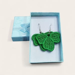 Op de afbeelding: Een paar groene, bladvormige oorbellen met zilveren haken ligt in een lichtblauwe geschenkdoos. De oorbellen zijn gemaakt van gedetailleerd, geborduurd materiaal. Een tweede, gesloten geschenkdoos met een blauw lint is zichtbaar.