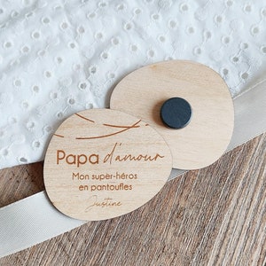 May include: Two wooden oval-shaped signs with a black magnet on the back. The top sign is blank. The bottom sign has the text "Papa d'amour" with the words "Mon super-héros en pantoufles" and "Justine" below.