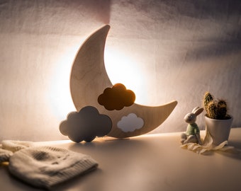 May include: A wooden crescent moon night light with three clouds, one brown, one gray, and one white. The light is on and illuminating the clouds. There is a small white ceramic bunny figurine and a cactus in a white pot on a white surface.