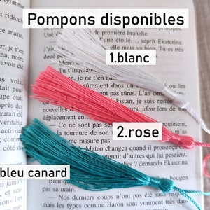 Puede incluir: Tres borlas de colores: blanco, rosa y verde azulado. La imagen incluye el texto en franc&eacute;s "Pompons disponibles" y los nombres de los colores "blanc", "rose" y "bleu canard". Las borlas se muestran sobre un libro abierto.