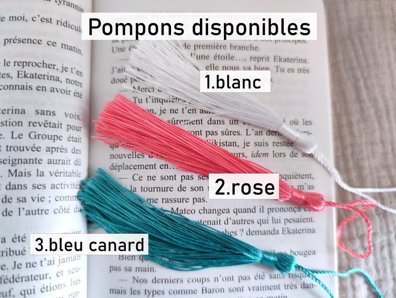 Puede incluir: Tres borlas de colores: blanco, rosa y verde azulado. La imagen incluye el texto en franc&eacute;s "Pompons disponibles" y los nombres de los colores "blanc", "rose" y "bleu canard". Las borlas se muestran sobre un libro abierto.