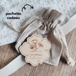 May include: A wooden flower-shaped tag with the name "Suzanne" and the text "Super ATSEM" engraved, next to a small beige drawstring bag. The tag features a floral design. The background includes a white ribbon and a white, eyelet fabric.