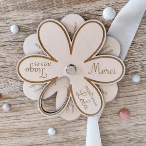 May include: A wooden flower-shaped craft with laser-cut petals. The center holds a silver fastener. The petals feature the words "Merci", "Tiago", and "Lola". A white ribbon and decorative beads are also present.