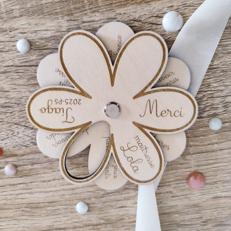 May include: A wooden flower-shaped craft with laser-cut petals. The center holds a silver fastener. The petals feature the words "Merci", "Tiago", and "Lola". A white ribbon and decorative beads are also present.