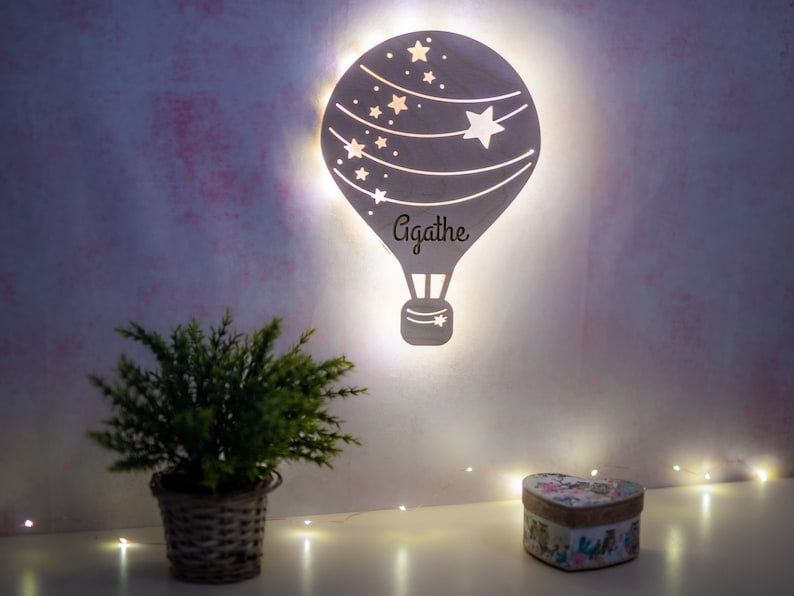 May include: A white wooden hot air balloon night light with a star design and the name Agathe inscribed on it. The light is illuminated and surrounded by white fairy lights.