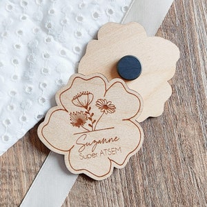 May include: Wooden flower-shaped magnet with engraved floral design and the name "Suzanne" and the text "Super ATSEM". The magnet has a dark gray circular magnet on the back. The background includes a white fabric and a wooden surface.