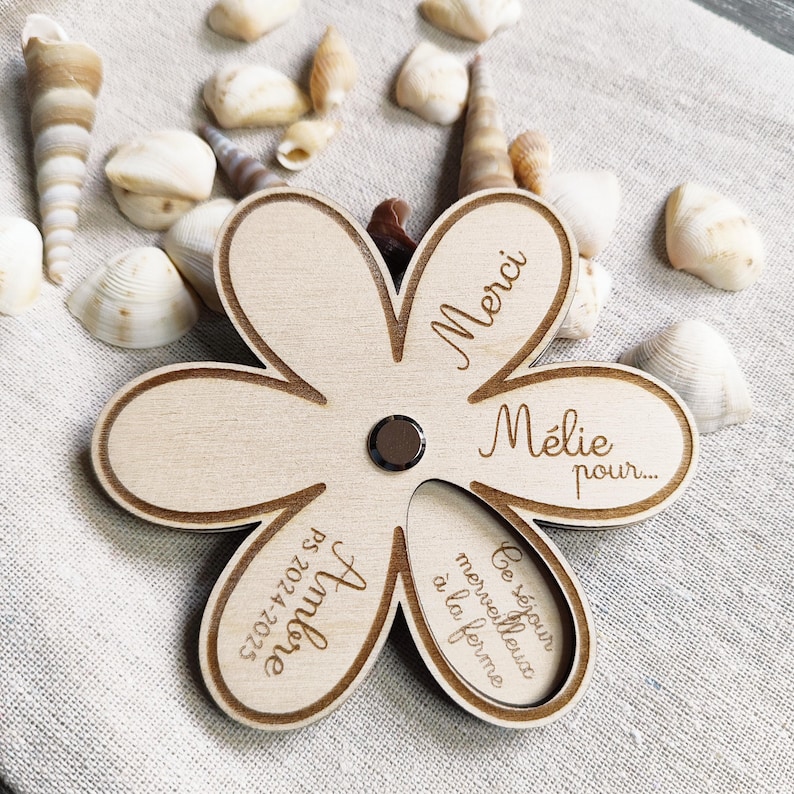 May include: A wooden flower-shaped magnet with engraved text. The magnet has a dark center and is surrounded by light-colored wood. The text includes the words "Merci", "M&eacute;lie pour...", "Ambre PS 2024-2025", and "Ce s&eacute;jour merveilleux &agrave; la ferme".