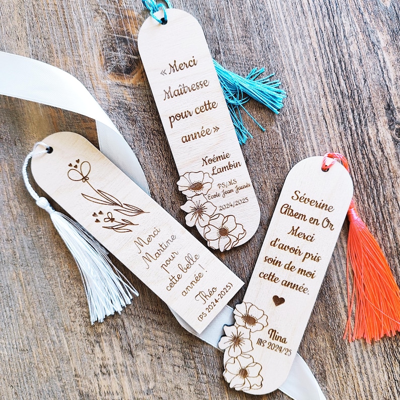 Puede incluir: Marcadores de libros de madera con borlas blancas, azules y naranjas. Cada marcador tiene un dise&ntilde;o &uacute;nico, con patrones florales y texto grabado en franc&eacute;s. El texto incluye nombres, fechas y frases como "Merci" y "Ma&icirc;tresse".