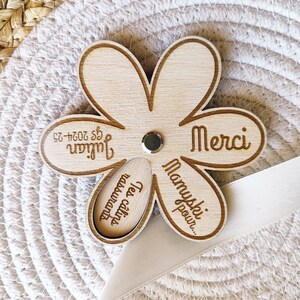 May include: A wooden flower-shaped keepsake with laser-etched text. The flower has five petals and a smaller, oval-shaped piece in the center. The text includes the name "Julian" and the word "Merci". A small, silver-colored stud is in the center.
