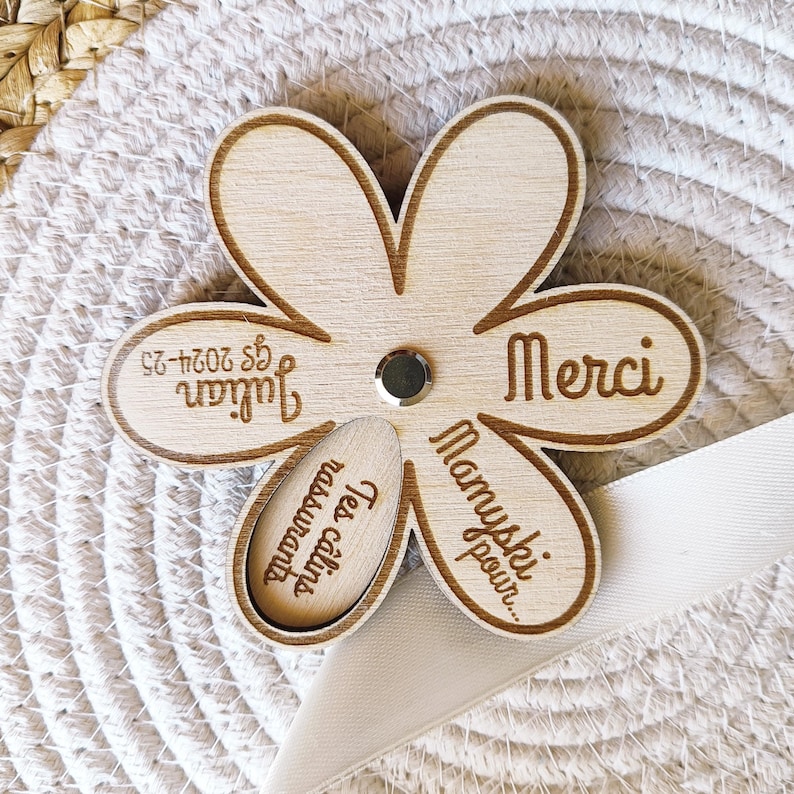 May include: A wooden flower-shaped keepsake with laser-etched text. The flower has five petals and a smaller, oval-shaped piece in the center. The text includes the name "Julian" and the word "Merci". A small, silver-colored stud is in the center.