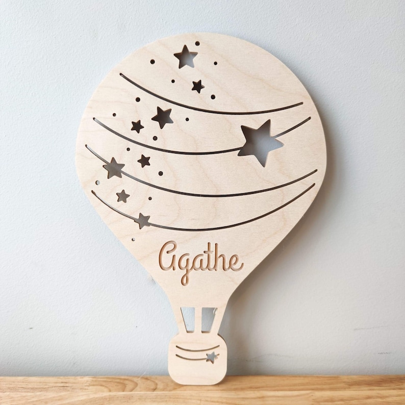 May include: A wooden hot air balloon cutout with a star pattern and the name Agathe inscribed on the bottom. The balloon is light brown and has a white background.