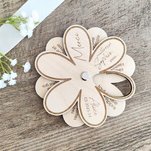 May include: A wooden flower-shaped gift with laser-etched text. The flower has multiple petals, with the central petal reading "Merci". The text "maitresse Sophie" and "Alice GS 2023-24" are also visible. A small silver magnet is in the center.
