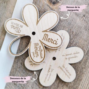 May include: Two wooden flower-shaped plaques with laser-etched text. One says "Merci" and "Nina MS 2024/25". The other says "M'avoir aid&eacute;e &agrave; grandir" and "ton sourire &agrave; toute &eacute;preuve". The plaques are light brown with a hole in the center.