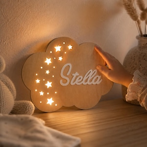 May include: A cloud-shaped wooden wall light featuring a warm glowing light emanating from star-shaped cutouts and small circular perforations. The name Stella is written in white, raised script lettering across the centre of the wooden surface.