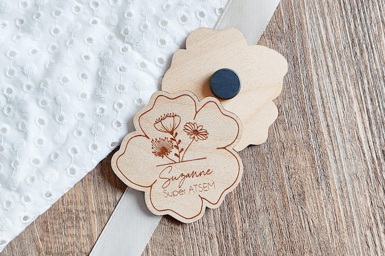 May include: Two wooden flower-shaped magnets. One magnet features an engraved floral design with the name "Suzanne" and the text "Super ATSEM". The other magnet has a black circular magnet attached to the back. The magnets are on a wooden surface.