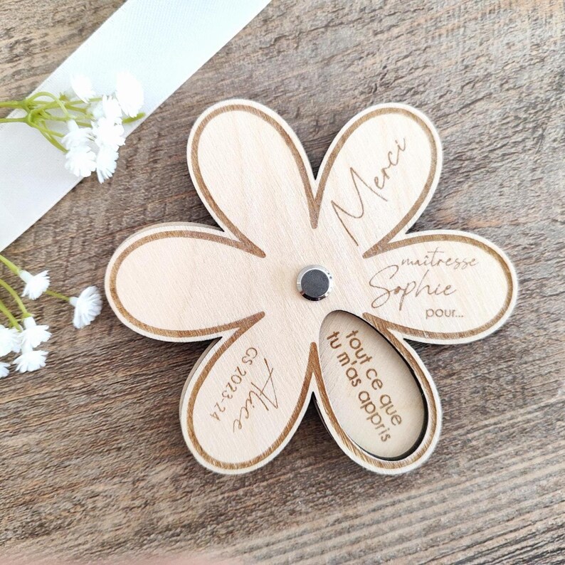 May include: A wooden flower-shaped magnet with a light brown finish. The magnet has six petals and features engraved text, including "Merci", "ma&icirc;tresse Sophie", and "Alice GS 2023-24". A small, round magnet is in the center.