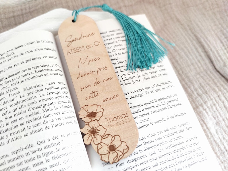 Puede incluir: Marcap&aacute;ginas de madera con texto grabado y dise&ntilde;o floral. El marcap&aacute;ginas tiene una borla turquesa y est&aacute; colocado en un libro abierto. El texto incluye nombres y un mensaje de agradecimiento.