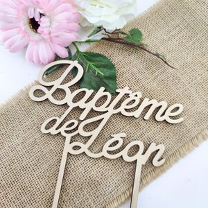 Peut inclure: Topper de g&acirc;teau en bois avec le texte "Bapteme de Leon" en &eacute;criture cursive. Le topper est sur un fond en toile de jute avec des fleurs roses et blanches.