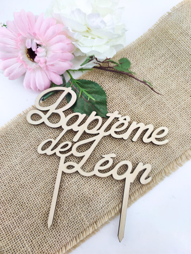 Peut inclure: Topper de g&acirc;teau en bois avec le texte "Bapteme de Leon" en &eacute;criture cursive. Le topper est sur un fond en toile de jute avec des fleurs roses et blanches.