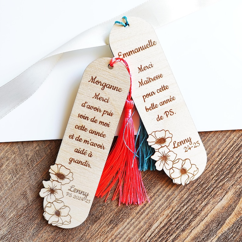 Puede incluir: Dos marcap&aacute;ginas de madera con dise&ntilde;os florales y borlas. Uno dice "Morganne Merci d'avoir pris soin de moi cette ann&eacute;e et de m'avoir aid&eacute; &agrave; grandir." El otro dice "Emmanuelle Merci Ma&icirc;tresse pour cette belle ann&eacute;e de PS."