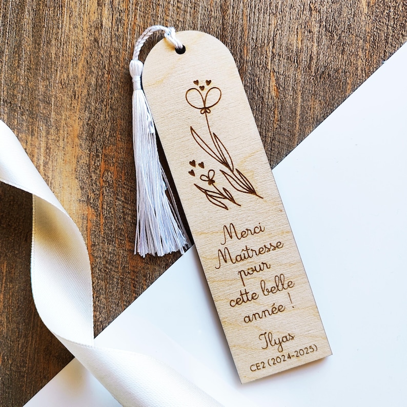 Puede incluir: Marcap&aacute;ginas de madera con dise&ntilde;o floral y borla blanca. El marcap&aacute;ginas tiene grabado el texto "Merci Ma&icirc;tresse pour cette belle ann&eacute;e !" y el nombre "Ilyas" y "CE2 (2024-2025)".