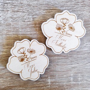 May include: Two wooden flower-shaped coasters with engraved floral designs and the names "Emma," "Merci Nathalie," and "Merci Julie." The coasters are light-colored with brown engraved details.