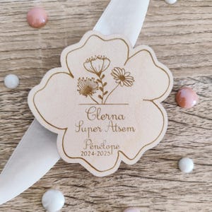 May include: A wooden, flower-shaped tag with engraved floral designs and the text "Clerna Super Atsem Penelope 2024-2025". The tag is light brown and rests on a white ribbon, surrounded by pink and white candies.
