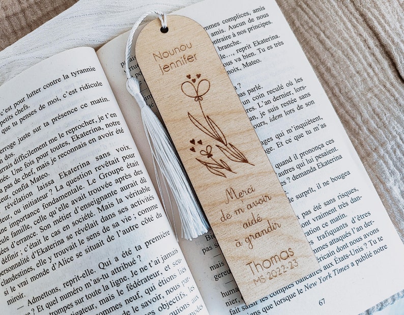 Puede incluir: Marcap&aacute;ginas de madera con borla blanca, grabado con un dise&ntilde;o floral y las palabras "Nounou Jennifer" y "Merci de m'avoir aid&eacute; &agrave; grandir". Incluye el nombre "Thomas" y el a&ntilde;o "MS 2022-23".