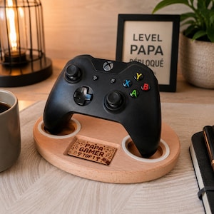 Può includere: Un controller Xbox nero è appoggiato su un supporto in legno con due aperture circolari e una piccola targa incisa con la scritta "PAPA GAMER TOP 1?". Un cartello incorniciato dietro il controller dice "LEVEL PAPA DÉBLOQUÉ".