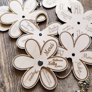 May include: Wooden flower-shaped tags with laser-etched text. The tags have a light wood color with darker brown edges and text. Some tags have the word "Merci" and names like "Nelly" and "Thyana".