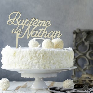 Peut inclure: G&acirc;teau blanc avec un topper de g&acirc;teau en bois qui dit "Bapt&ecirc;me de Nathan". Le g&acirc;teau est d&eacute;cor&eacute; de gla&ccedil;age blanc et de copeaux de chocolat blanc.