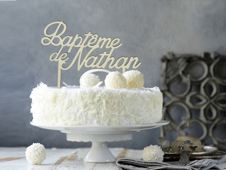 Peut inclure: G&acirc;teau blanc avec un topper de g&acirc;teau en bois qui dit "Bapt&ecirc;me de Nathan". Le g&acirc;teau est d&eacute;cor&eacute; de gla&ccedil;age blanc et de copeaux de chocolat blanc.