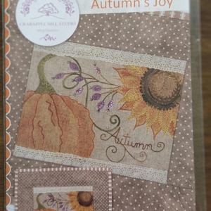 May include: A package featuring an autumn-themed embroidery design. The design includes a pumpkin, sunflower, and the word "Autumn" in script. The background is a neutral color with a dotted pattern, and the text "Autumn's Joy" is visible.