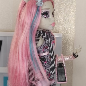 May include: A doll with long pink hair featuring blue streaks, pale grey skin, and pink lips. The doll wears a black and white patterned top with pink accents, a pink ruffled skirt, and a black handbag with pink trim. A silver fleur-de-lis hair accessory is visible.