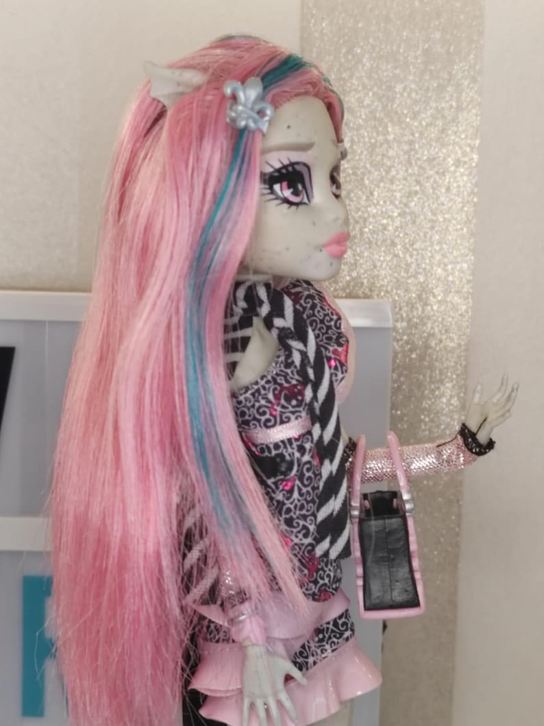 May include: A doll with long pink hair featuring blue streaks, pale grey skin, and pink lips. The doll wears a black and white patterned top with pink accents, a pink ruffled skirt, and a black handbag with pink trim. A silver fleur-de-lis hair accessory is visible.