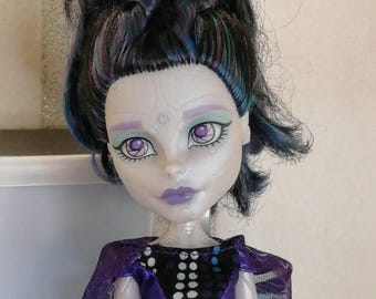 以下が含まれることがあります： 青い花を髪につけた、紫と黒のドレスを着た人形。白い肌に紫色のアイシャドウ、紫と黒の衣装を着ています。