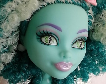 May include: A doll with teal skin, curly teal and white hair, and a small pink hat. The doll has large green eyes with black pupils, purple eyeshadow, and glossy light purple lips. It wears a light purple top with a black lace collar detail.