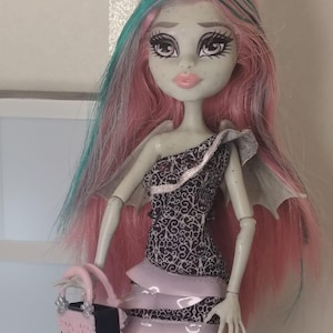 May include: A doll with pale grey skin, freckles, and long pink hair with teal streaks. The doll wears a one-shoulder dress featuring a black and white ornate pattern with layered light pink ruffles and carries a small pink and black handbag.
