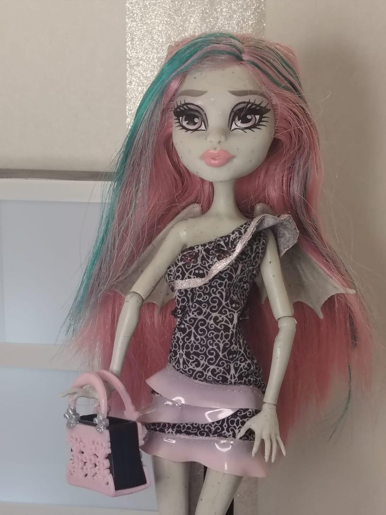 May include: A doll with pale grey skin, freckles, and long pink hair with teal streaks. The doll wears a one-shoulder dress featuring a black and white ornate pattern with layered light pink ruffles and carries a small pink and black handbag.