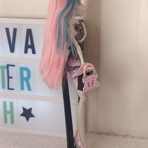 May include: A doll with long pink hair featuring blue streaks, wearing a patterned top and skirt. The doll holds a small pink and black handbag with cutout details and wears silver and pink platform shoes, all supported by a black stand.