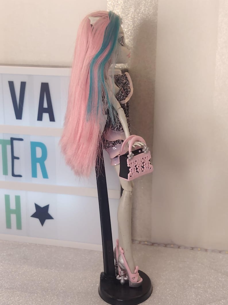 May include: A doll with long pink hair featuring blue streaks, wearing a patterned top and skirt. The doll holds a small pink and black handbag with cutout details and wears silver and pink platform shoes, all supported by a black stand.