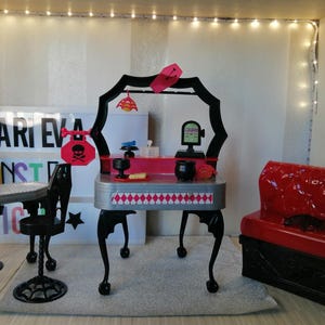 Peut inclure: Un ensemble de jeu de maison de poupée noir et rouge avec une coiffeuse, deux chaises et un canapé. La coiffeuse a un motif à carreaux noir et blanc et une enseigne avec un crâne et des os croisés. L'ensemble est décoré d'accents roses et noirs.
