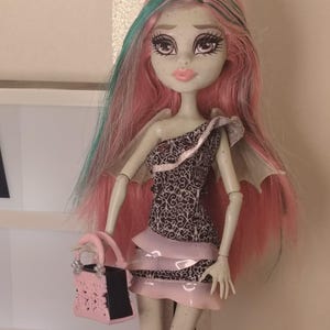 May include: A doll with pale skin and long pink hair featuring teal streaks. The doll wears a one-shoulder dress with a black and white filigree pattern and layered pink ruffles. It has small wings on its back and holds a pink and black handbag.