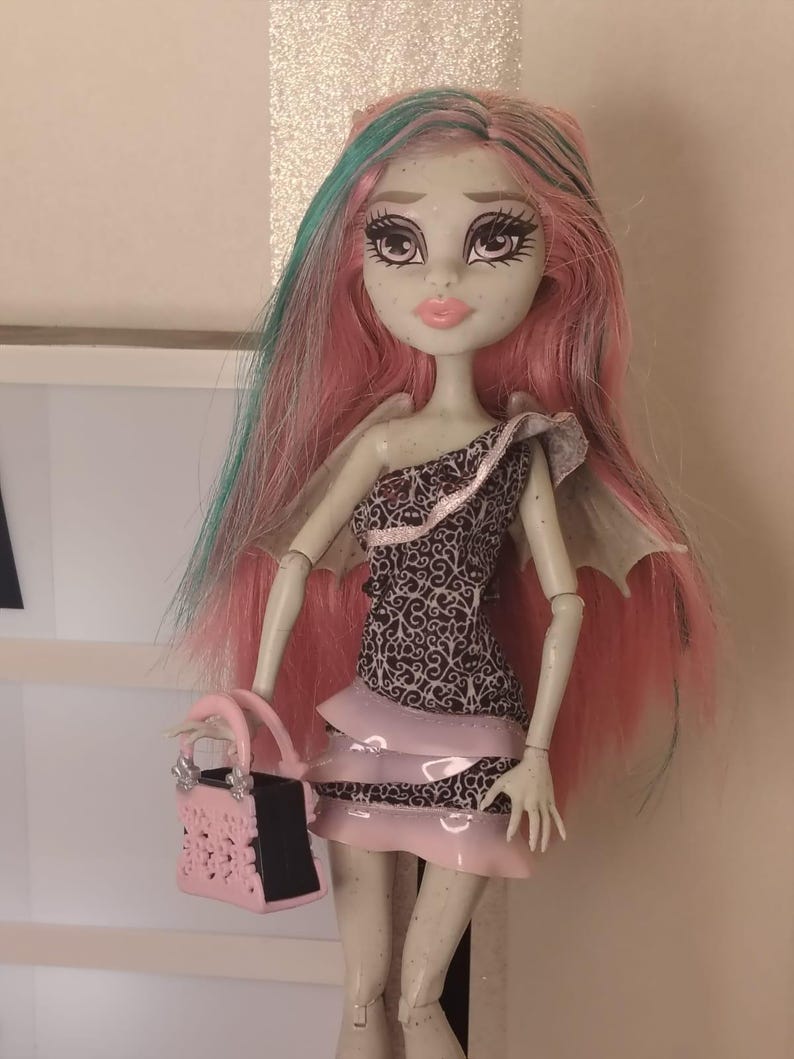 May include: A doll with pale skin and long pink hair featuring teal streaks. The doll wears a one-shoulder dress with a black and white filigree pattern and layered pink ruffles. It has small wings on its back and holds a pink and black handbag.