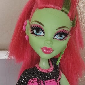 Puede incluir: Muñeca con piel verde, cabello rosa brillante y ojos azules. La muñeca tiene labios rosas y lleva una blusa negra y rosa. La muñeca tiene pendientes rosas y un detalle verde en forma de vid en el costado de la cara.