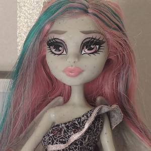 May include: A doll with light grey skin, freckles, and long pink hair with teal streaks. The doll has large pink eyes with dark eyelashes, light pink lips, and small grey wings on the back. It wears a black dress with a white intricate pattern.