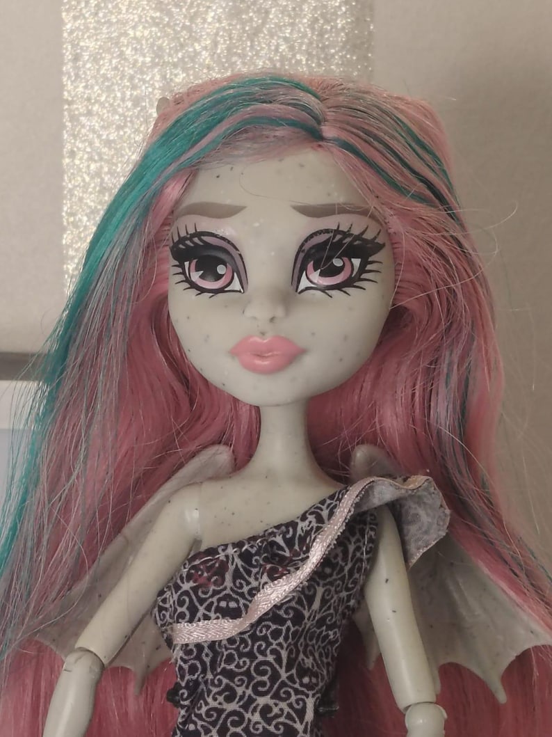 May include: A doll with light grey skin, freckles, and long pink hair with teal streaks. The doll has large pink eyes with dark eyelashes, light pink lips, and small grey wings on the back. It wears a black dress with a white intricate pattern.