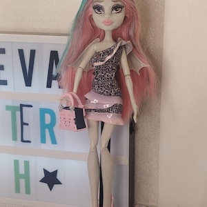 May include: A doll with pale skin, freckles, and long pink hair with teal streaks. It wears a one-shoulder dress with a black and white filigree pattern and pink ruffled trim, small wings, pink platform heels, and carries a small pink handbag.