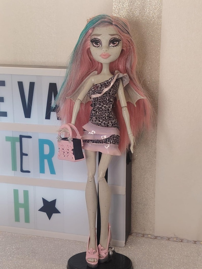 May include: A doll with pale skin, freckles, and long pink hair with teal streaks. It wears a one-shoulder dress with a black and white filigree pattern and pink ruffled trim, small wings, pink platform heels, and carries a small pink handbag.